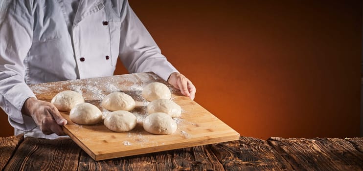 Chef in a pizzeria with tray of raw dough in individual portions for the pizza crust over a brown background with copy space in a wide angle panorama with glow of the oven fire