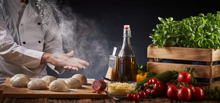 Chef preparing a speciality homemade Italian pizza clapping his hands in a cloud of flour above portions of raw dough on a table with assorted fresh ingredients in a panorama banner
