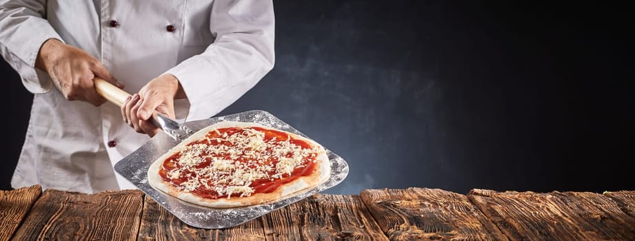 Chef making a traditional Italian pizza holding the crust with tomato paste and grated cheese on a paddle over a rustic table in a panorama banner with copy space