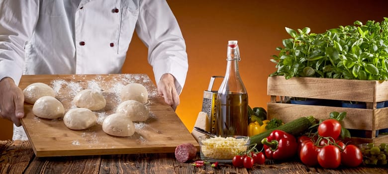Chef preparing fresh raw dough for pizzas in a pizzeria carrying a board with individual portions to a table with fresh topping ingredients in a panorama banner