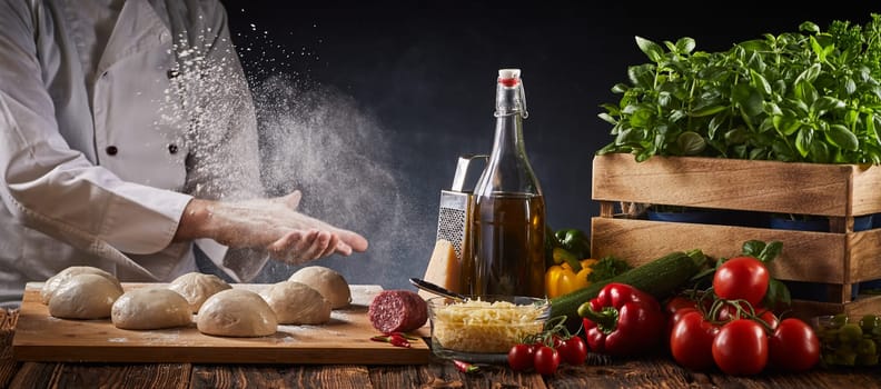 Chef clapping his floured hands creating a cloud of flour in the air as he prepares dough portions for pizzas on a table with fresh assorted ingredients in a panorama banner