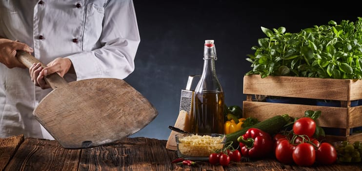 Chef holding a wooden paddle in a pizzeria above a rustic table with assorted fresh ingredients for Italian cuisine in a panorama banner