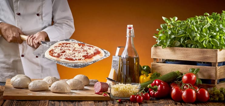 Cook preparing an Italian pizza in a restaurant holding a paddle with pastry base behind a rustic table with assorted fresh ingredients and portions of dough in a panorama banner