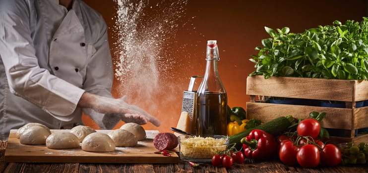 Freeze motion explosion of cooking flour as a chef preparing pizzas in a pizzeria claps his hands over portions of raw dough on a tray alongside fresh ingredients in a panorama banner