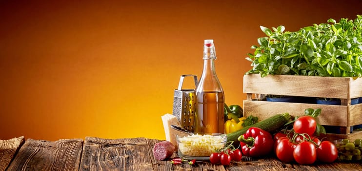 Ingredients for Italian cuisine in a pizzeria with parmesan cheese, olive oil, tomatoes, basil and sweet peppers on a rustic table over a brown background with copy space in a wide angle panorama with glow of the oven fire