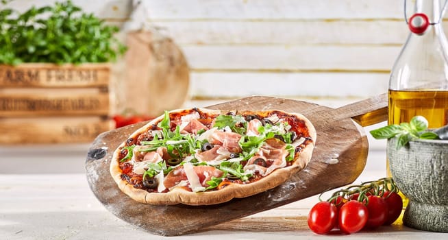 Thin crust Italian pizza with prosciutto ham, fresh rocket leaves, parmesan cheese flakes and olives on a wooden paddle with ingredients in a pizzeria suitable for menu advertising