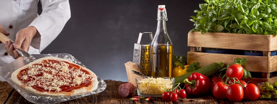Chef making a traditional Margherita pizza holding a paddle with crusty base, tomato and grated cheese above a table with fresh ingredients in a panorama banner