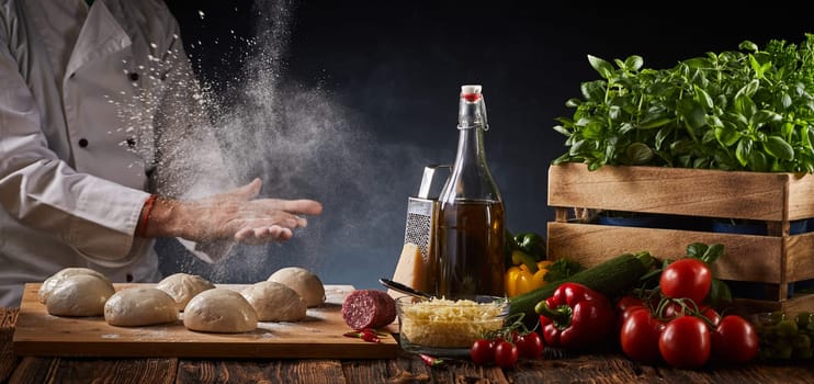 Chef clapping his hands with suspended flour in a freeze motion view as he places portions of raw dough alongside fresh pizza ingredients on a rustic table