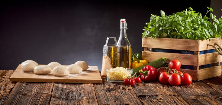 Panorama banner with fresh pizza ingredients, grated cheese, olive oil and mounds of individual portions of dough on a cutting board on a rustic kitchen table over a dark background with copy space