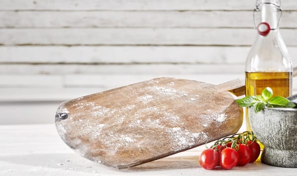 Empty floured wooden pizzeria paddle with ripe red vine tomatoes, olive oil and basil in a pestle and mortar with copy space
