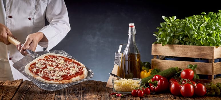 Chef making a traditional Margherita pizza holding a paddle with pie crust covered in tomato paste and cheese over a table with assorted fresh ingredients in a panorama banner