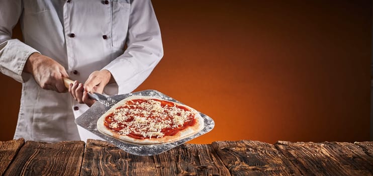 Chef in a pizzeria preparing an Italian pizza holding a crusty base with tomato paste and grated cheese on a metal paddle against a rustic table over a brown background with copy space in a wide angle panorama with glow of the oven fire