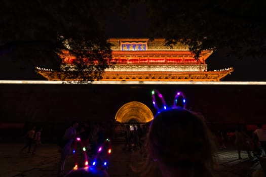 Xian, China -  July 2019 : Children with lighting LED ears accesory on their heads standing in front of the Xian Bell Drum Tower beautifully lit and illuminated at night, Shaaxi Province