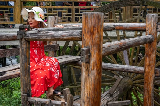 Wullingyuan, China -  August 2019 : Female dressed in a red dress using her legs to pull levers on a wheel of an old Water wheel in Huanglong Yellow Dragon Cave scenic area, Hunan province