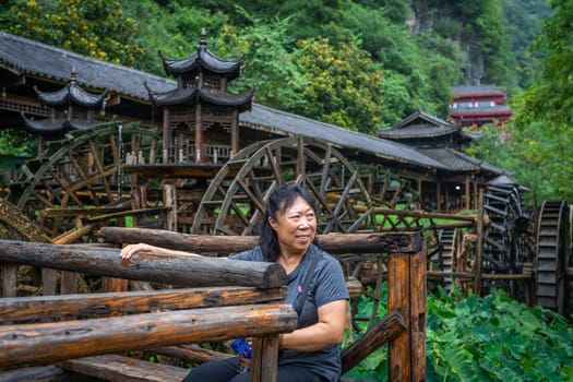 Wullingyuan, China -  July 2019 : Older chinese woman using her legs to pull levers on a wheel of an old Water wheel in Huanglong Yellow Dragon Cave scenic area, Zhangjiajie, Hunan Province