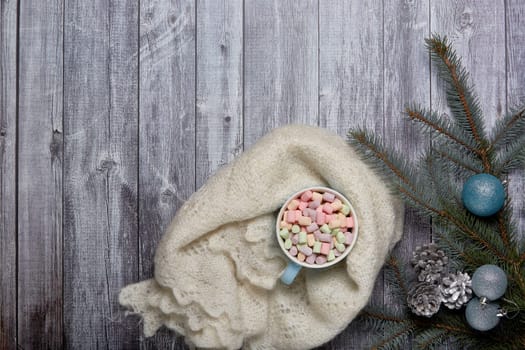 Mug of cocoa with marshmallow, woolen downy shawl, fir tree branches and Christmas ornament on grey wooden background. Top view with copy space. New Year and Christmas holiday