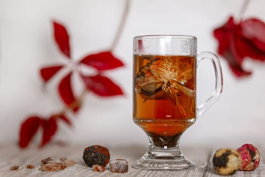 Open flower Chinese tea in a glass on a wooden stand against a background of red leaves and balls of dry tea and caramelized sugar near a glass