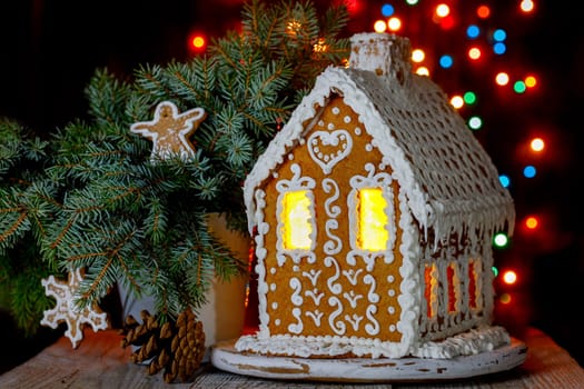 Gingerbread house with bokeh of light bulbs in the background and sprigs of blue spruce