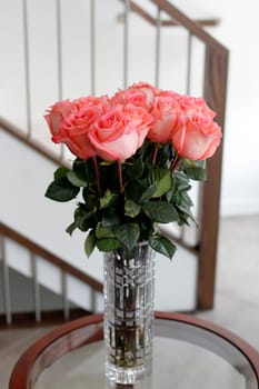 Beautiful darker pink rose flowers with dark green leaves in a glass crystal vase on a living room side table. Pink roses flower bouquet in a clear tall crystal vase on a round table near a stairwell.