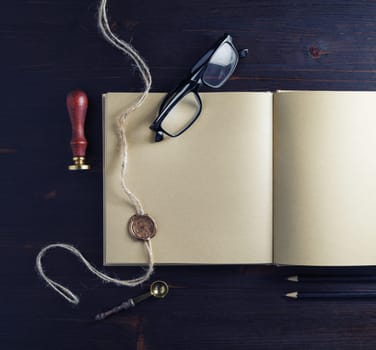 Opened kraft paper brochure and stationery on wood table background. Book, glasses, stamp, sealing wax and pencils. Flat lay.