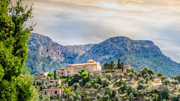 Beautiful old town of Deia with church of Saint Joan Baptista on terraced hill with high mountains and cliffs in background, sunset, Mallorca, Spain