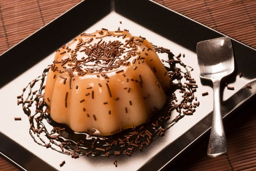 Caramel pudding dessert decorated with sweet sauce and chocolate crumbs on black square plate.