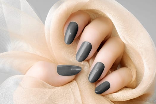 Female hand with dark gray nails is holding beige textile, nail care and manicure concept.