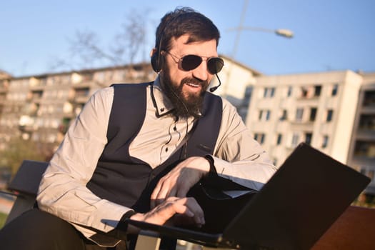 The middle aged bearded smiling man is working at virtual assistent freelance work on his laptop outdoors in city at the street.