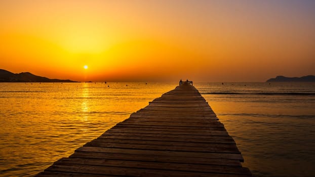 Orange solar disc in sunrise on beach with silhouette of mountains and pier on bay, reach orange and yellow colours of the sky panorama Mallorca Spain