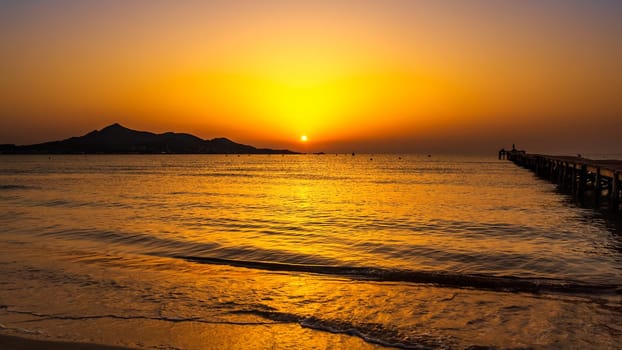 Orange solar disc in sunrise on beach with silhouette of mountains and pier on bay, reach orange and yellow colours of the sky panorama Mallorca Spain