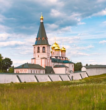 Monastery with bell tower and golden church domes. Meadow and wall in foreground with blue cloudy sky in background