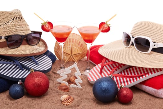 Beach accessories (flip flops, straw hats, seashells, sand, sunglasses), two glasses of cocktail, christmas ornaments on white background. Christmas. Winter vacation in warm countries, beach holiday