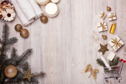 Shopping trolley with gift boxes, spa composition with cosmetic creams, towel and Christmas ornaments on light wooden background. Top view with copy space. Christmas and New Year sale.