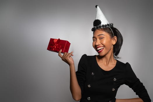 Pretty happy Asian girl in birthday cap holding present isolated over gray wall background