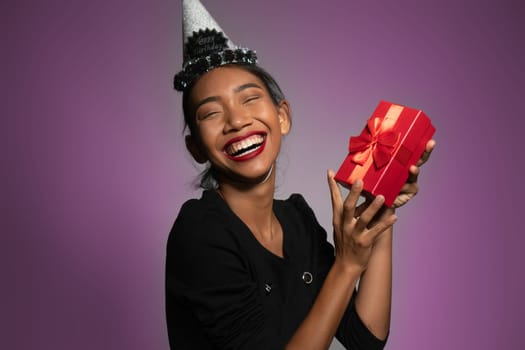 Pretty happy Asian girl in birthday cap holding present isolated over purple wall background