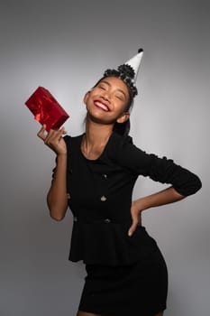 Pretty happy Asian girl in birthday cap holding present isolated over gray wall background