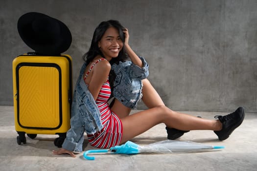 Portrait of smiling young Asian woman with luggage isolated over concrete wall background. People lifestyle, travel concept