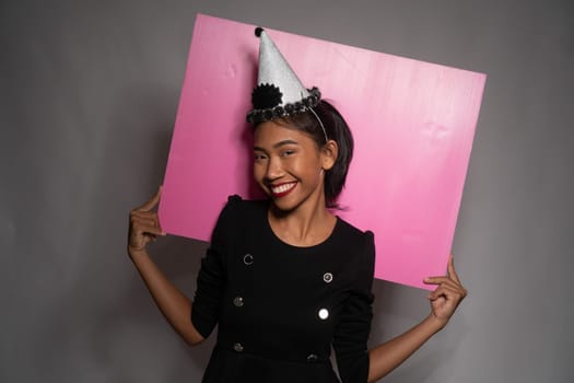 Pretty happy Asian girl in birthday cap holding pink board behind her isolated over gray wall background