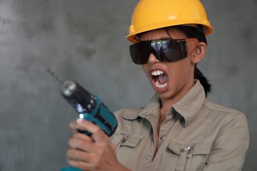 Portrait of attractive young girl builder with drill in overall, helmet and protective glasses isolated over concrete wall background