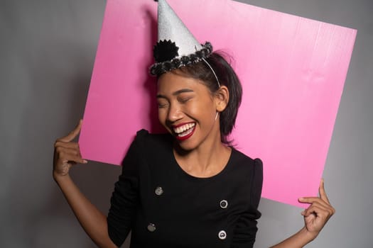 Pretty happy Asian girl in birthday cap holding pink board behind her isolated over gray wall background