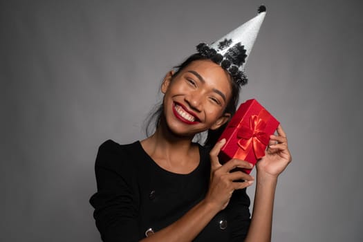 Pretty happy Asian girl in birthday cap holding present isolated over gray wall background