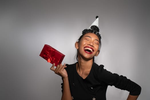 Pretty happy Asian girl in birthday cap holding present isolated over gray wall background