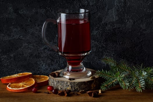 Mulled wine in a glass cup on a wooden platter, on the background of a fir branch and dry oranges