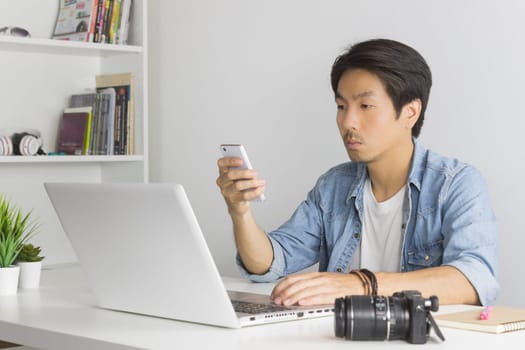Asian Photographer or Freelancer in Denim or Jeans Shirt Send Message to Customer by Smartphone in front of Laptop in Home Office. Photographer or freelancer working with technology