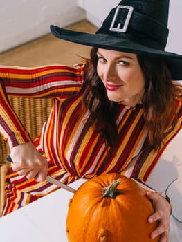 Beautiful young woman wearing witch's black hat  in her kitchen cutting a pumpkin for Halloween.