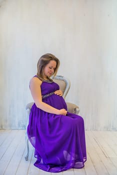 Beautiful pregnant woman in ultra violet dress sitting on the armchair