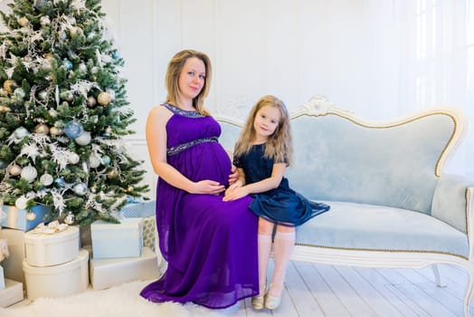 Beautiful pregnant woman in ultra violet dress sitting with daughter on the sofa near christmas tree