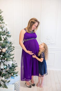 Beautiful pregnant woman in ultra violet dress standing with daughter on the sofa near christmas tree
