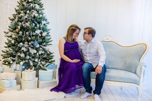 Pregnant woman in ultra violet dress with husband sitting on the sofa near christmas tree