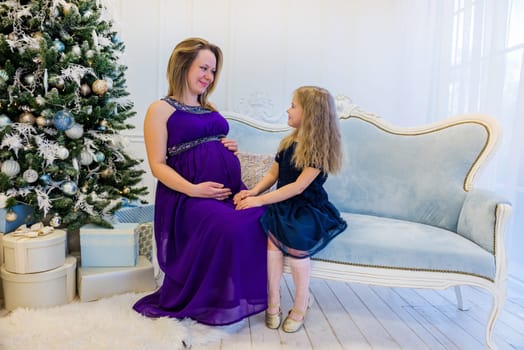 Beautiful pregnant woman in ultra violet dress sitting with daughter on the sofa near christmas tree